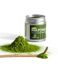 A jar of Arteamony Bio Matcha ceremonial grade green tea powder, shown with a small pile of vibrant green powder, a wooden spoon with loose matcha, and a clean white background.