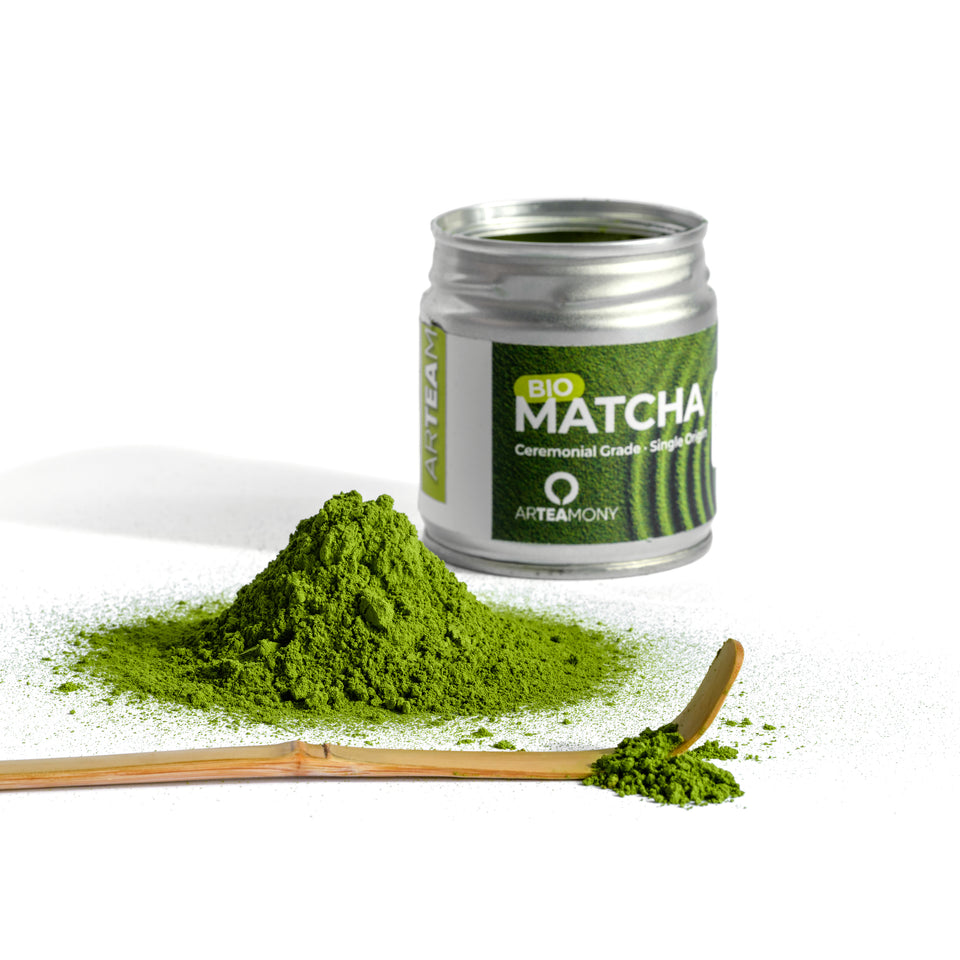 A jar of Arteamony Bio Matcha ceremonial grade green tea powder, shown with a small pile of vibrant green powder, a wooden spoon with loose matcha, and a clean white background.