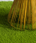 Close-up of a bamboo rake with thin golden-yellow tines resting on bright green grass against a light neutral background.