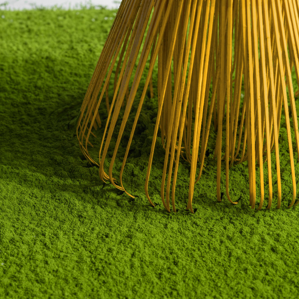 Close-up of a bamboo rake with thin golden-yellow tines resting on bright green grass against a light neutral background.