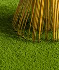Close-up of a bamboo rake with thin golden-yellow tines resting on bright green grass against a light neutral background.