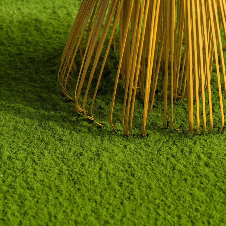 Close-up of a bamboo rake with thin golden-yellow tines resting on bright green grass against a light neutral background.
