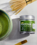 A styled flat-lay of a matcha tea product on a light marble surface, featuring a small silver canister labeled 'Matcha' with green swirl graphics, a bowl of prepared green tea, a bamboo whisk, and a bamboo scoop.