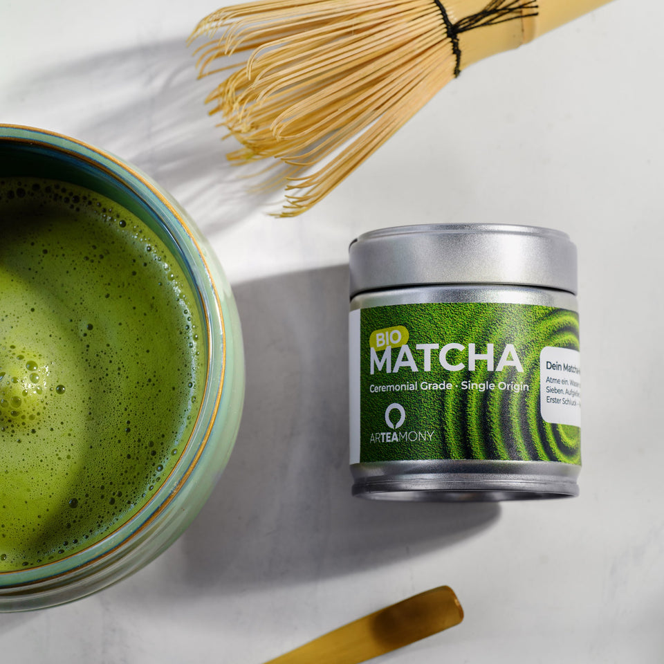 A styled flat-lay of a matcha tea product on a light marble surface, featuring a small silver canister labeled 'Matcha' with green swirl graphics, a bowl of prepared green tea, a bamboo whisk, and a bamboo scoop.
