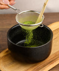 A close-up of hands using a small metal sieve with a bamboo handle to sift bright green matcha powder into a black ceramic bowl placed on a wooden board.