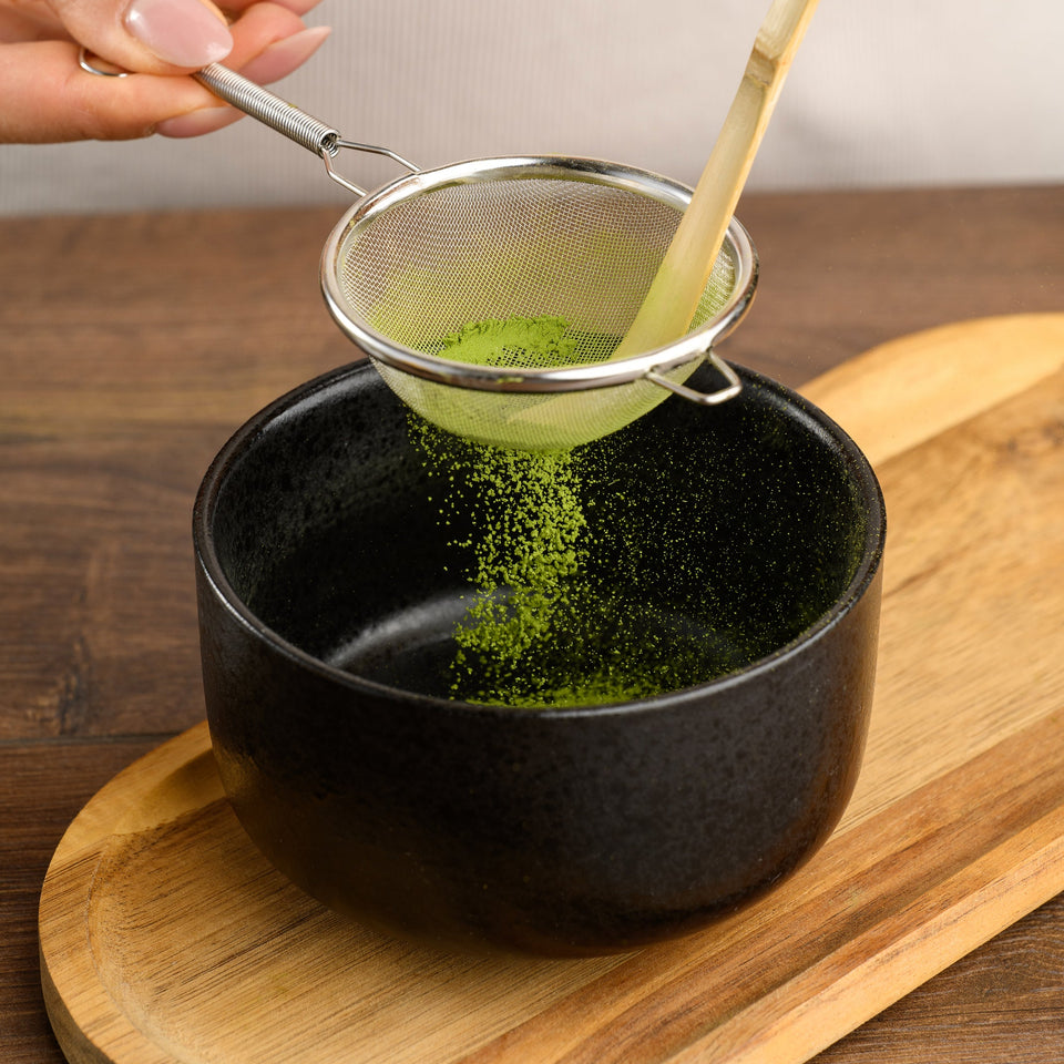 A close-up of hands using a small metal sieve with a bamboo handle to sift bright green matcha powder into a black ceramic bowl placed on a wooden board.