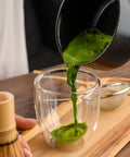 Green matcha tea being poured from a black ceramic bowl into a clear double-walled glass on a wooden tray, with a bamboo whisk and metal strainer nearby.