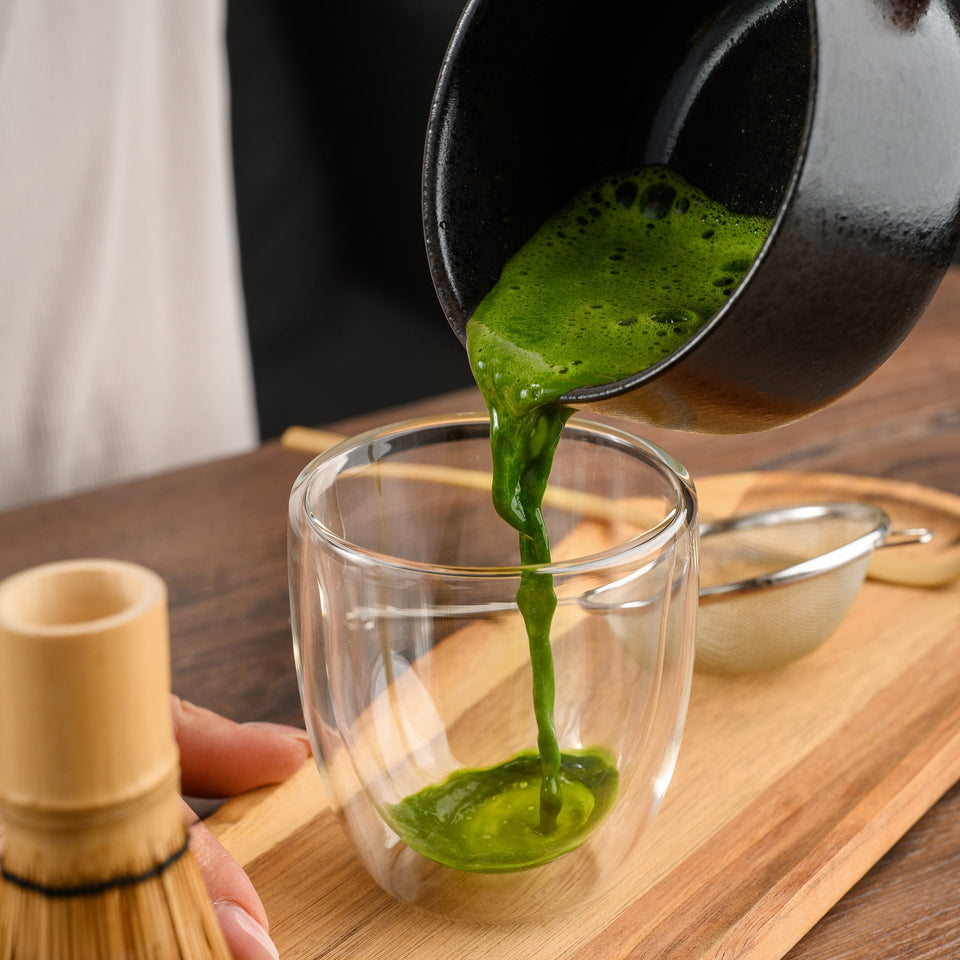 Green matcha tea being poured from a black ceramic bowl into a clear double-walled glass on a wooden tray, with a bamboo whisk and metal strainer nearby.