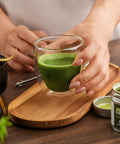 A person holds a clear double-walled glass cup filled with bright green matcha tea on a wooden tray, with a small jar of matcha powder nearby on a dark wood table.