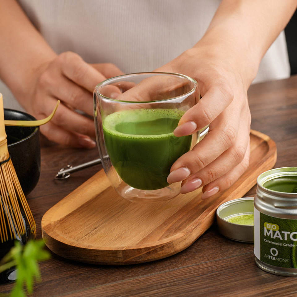 A person holds a clear double-walled glass cup filled with bright green matcha tea on a wooden tray, with a small jar of matcha powder nearby on a dark wood table.